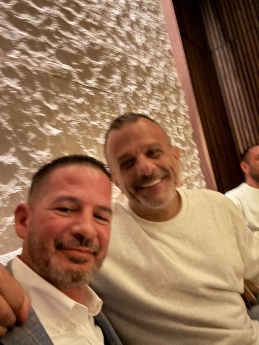 A selfie of two smiling middle-aged men with short hair and beards, posing together in front of a textured white stone wall.