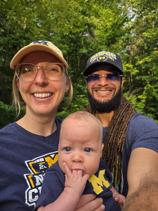 Outdoor selfie of two adults and a baby, with the woman wearing a Michigan cap and shirt, and the man wearing a 'CHAMPIONS' cap and sunglasses.