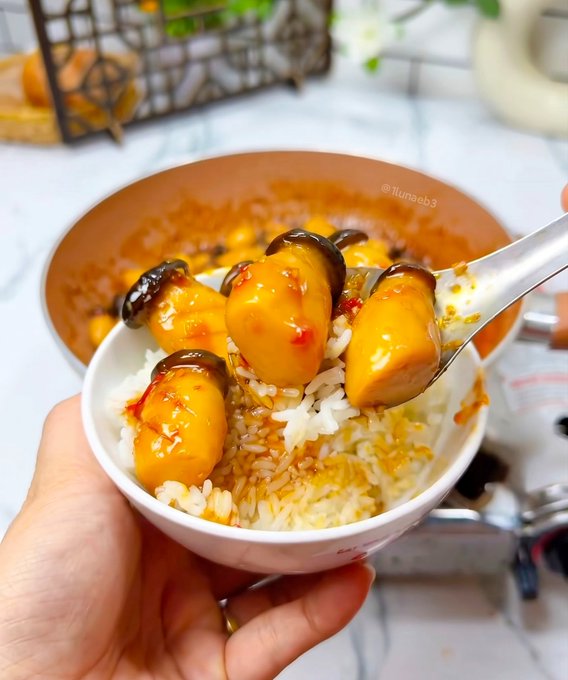 A hand holds a small bowl of rice topped with glazed mushrooms and orange-colored sauce, with a spoon lifting a portion, watermarked '@llunab3'.