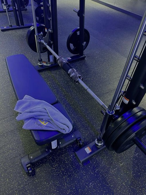 A photograph of gym equipment, specifically a weight bench with a blue towel draped over it, next to a barbell loaded with weight plates on a Smith machine or rack system.