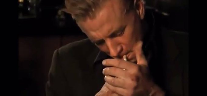A close-up shot of a man with blond hair in a dark suit, appearing deep in thought with his hands clasped near his face in a dimly lit setting.