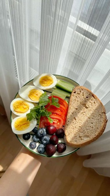 A plate held by a hand contains a breakfast meal of hard-boiled egg halves, sliced tomatoes, cucumber, fresh greens, blueberries, dark berries, and a large piece of bread, set against a window with sheer curtains.