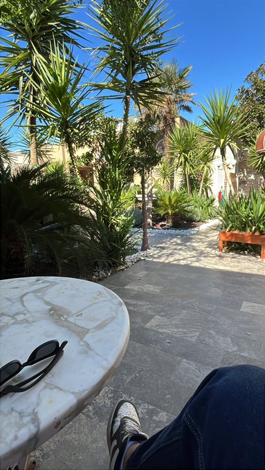 A POV shot from someone relaxing outdoors on a sunny day, showing their sneakers resting near a round marble table with sunglasses on it, overlooking a tropical garden with palm trees, yucca plants, and stone pathways.