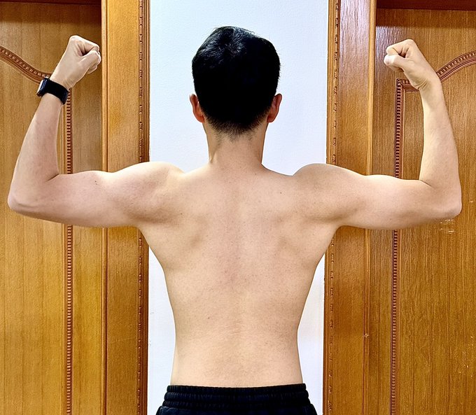 A shirtless man stands with his back to the camera, flexing both arms in a double bicep pose between two wooden doors, showing off a lean, defined physique. The image visually reinforces the post's message about the "crypto circle" favoring lean muscle, suggesting that fitness training is the price of entry for top-tier crypto culture.