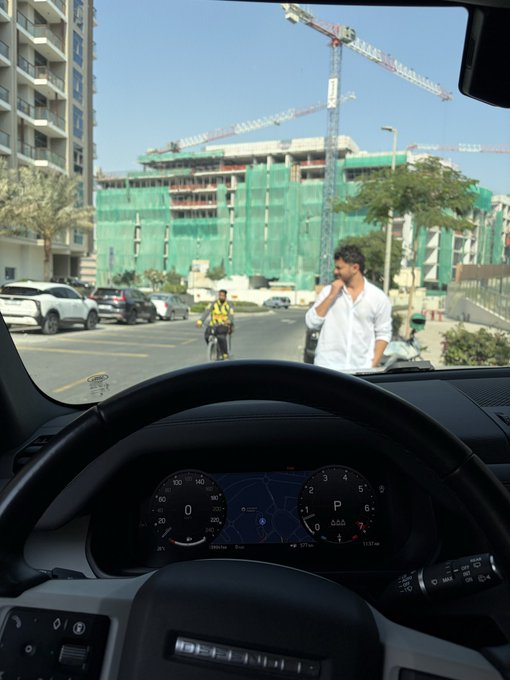 A view from the driver's seat of a vehicle (in Park), looking out through the windshield at a pedestrian in a white shirt walking across a street, with a construction site and crane visible in the background.