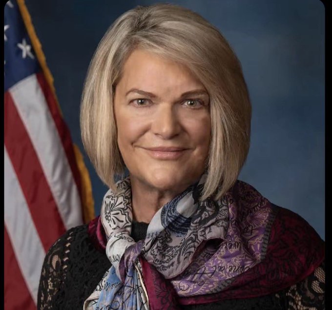 An official portrait of Senator Cynthia Lummis (R-WY), featuring a blonde woman smiling, wearing a patterned purple scarf and black top, with the American flag visible in the background.