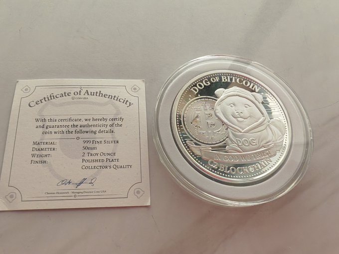 The image shows a silver coin labeled "DOG OFBITCOIN" and has an authenticity certificate attached, indicating that it is genuine. The coin has been received today by Cryptovation and C2Blockchain. The coin's size and cleanliness are of high quality and meet the standards for a real, valuable coin. It is presented in such a way that conveys its value, highlighting that it is a certified authentic item.