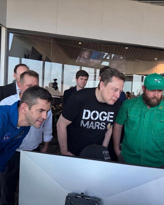 urn of water, a man in a black t-shirt with "DOGE MARS" printed on it, and another man in a green shirt stand before a large screen displaying the word "DOGE".