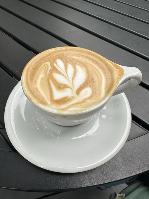 A cappuccino with leaf latte art sits in a white ceramic cup on a saucer atop a dark slatted café table. The image complements the post's message of savoring a calm, leisurely moment—reflecting the freedom that comes from long-term investing rather than the stress of constant trading.