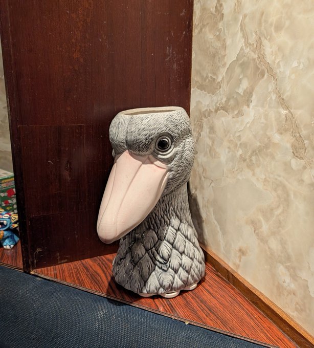 A gray ceramic dodo bird vase/figurine sits on a wooden floor in the corner of a room with marble-patterned wallpaper and dark wood paneling. The quirky decorative piece, with its distinctive large pink beak and textured feathered body, adds an unusual touch to the space where the singer met a friend for microtonal karaoke.