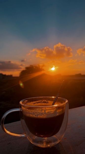A clear glass cup of dark coffee with a spoon sits on a ledge, silhouetted against a vibrant orange and yellow sunset.