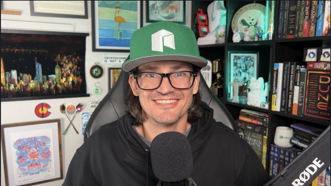 A man with glasses and a green cap sitting in front of a microphone, with shelves of collectibles, books, and framed art behind him.