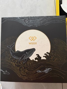 A black package or box from WEEX featuring an intricate illustration of a whale and dolphins swimming in golden-lined waves around a large sun or moon with the WEEX logo.