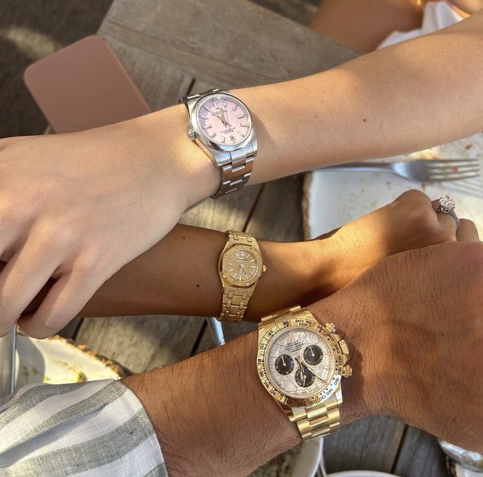 The image shows three arms resting together on a wooden table, each displaying an expensive luxury timepiece—a pink-dial Rolex with "OYSTER PERPETUAL DATE" visible, a gold Audemars Piguet, and a gold Rolex Cosmograph Daytona with "ROLEX" and "DAYTONA" text readable on the dial. The watches visually contrast with the post's message, which mocks people who equate material wealth (symbolized by the Rolex and AP) with success, suggesting that true intellectual substance—like debating $XRP cryptocurrency—reveals more about one's character than flashy possessions do.