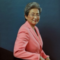 An older woman with short dark hair and glasses, wearing a pink blazer, smiles warmly against a dark blue background.