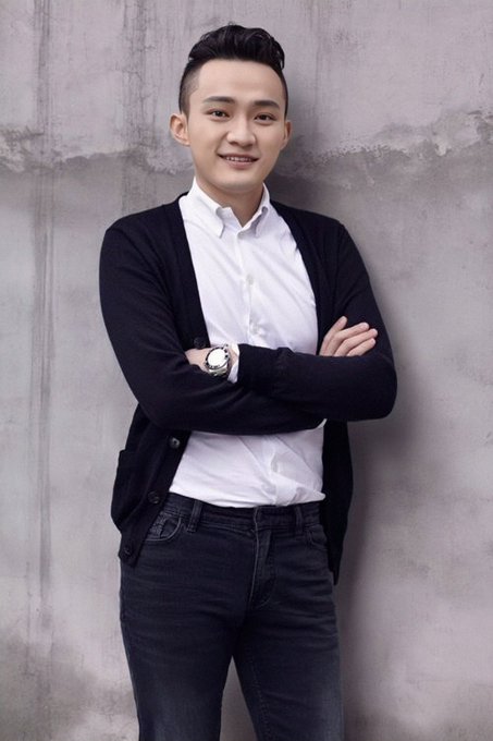 A young man smiles while posing against a gray concrete wall, wearing a white button-up shirt, black cardigan, jeans, and a wristwatch.