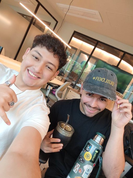 The image features two men sitting next to each other at a desk with their arms around each other's shoulders. One of them has a black hat, while the other one wears a white shirt. They are both holding drinks - specifically, cups filled with coffee or tea. The background includes a TV screen and some chairs placed in front of it. There is also a backpack on the floor next to one of the men.