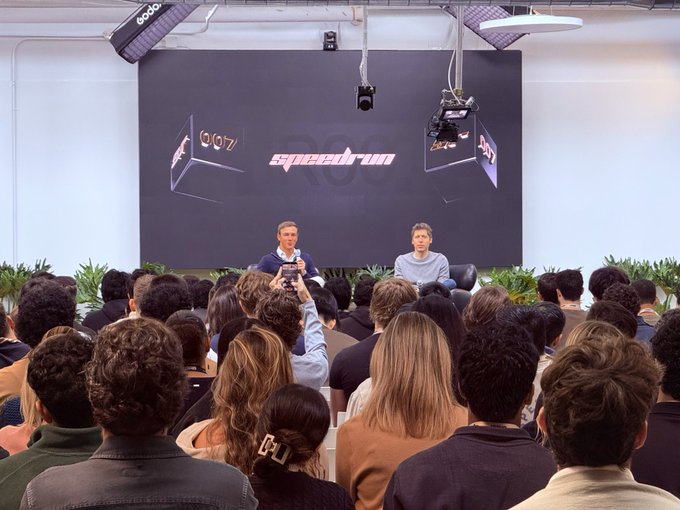 Two men sit on a stage in front of a 'speedrun' screen at an event, speaking to an audience as someone in the crowd records on a phone.