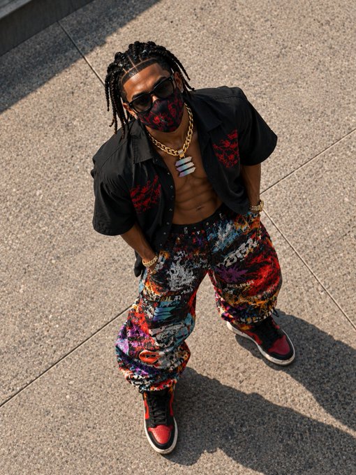A stylish man poses for a high-angle portrait, wearing an open black shirt, gold chains, sunglasses, a patterned face mask, colorful graffiti-style pants, and red, black, and white sneakers on a paved surface.
