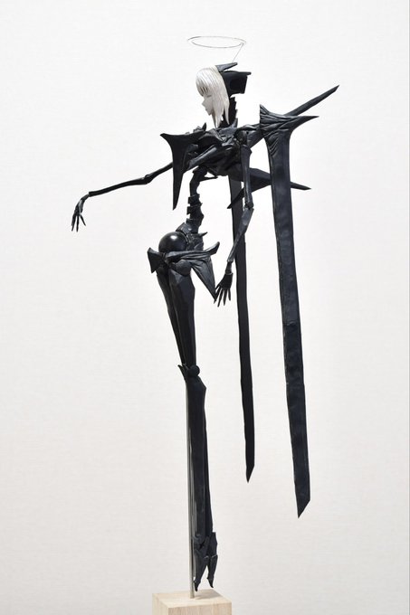 The image features a black metal sculpture of a woman with white hair, standing upright on her hind legs against a white background. The sculpture has long arms extending outwards from its body, giving it an ethereal appearance. The sculpture appears to be made of metal and is placed on a wooden base.
