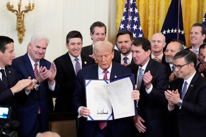 Former President Donald Trump holds up a document surrounded by men in suits, likely representing the administration and industry leaders. The image relates to the post by suggesting a historical context of high-level government engagement with business and policy.