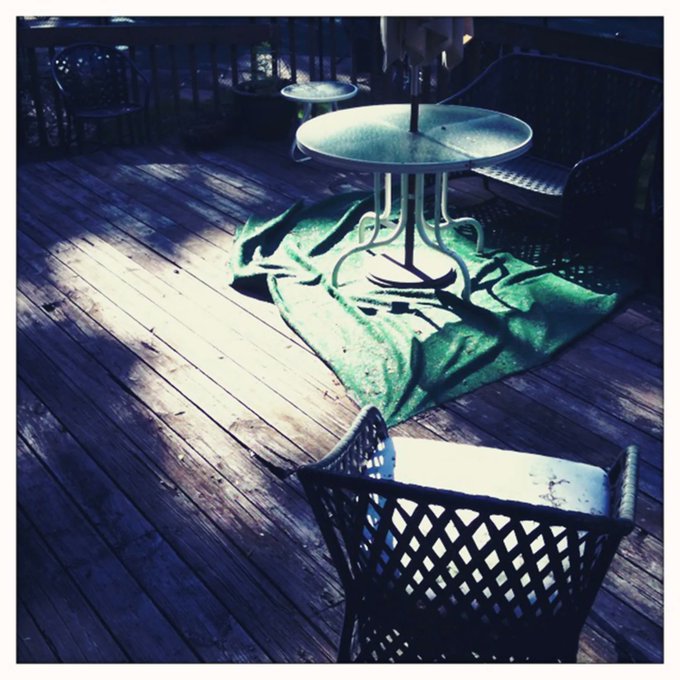 An atmospheric outdoor cafe setting at night, showing a wooden deck with metal tables, chairs, and a green cloth illuminated by warm light.
