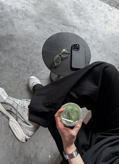 A first-person view of someone wearing black pants and white ASICS sneakers, holding a green matcha drink, with a phone and glasses resting on a round table nearby.
