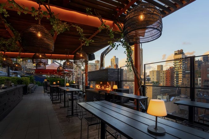 An outdoor rooftop bar at sunset with woven pendant lights, a fireplace, high-top tables, and a city skyline in the background.