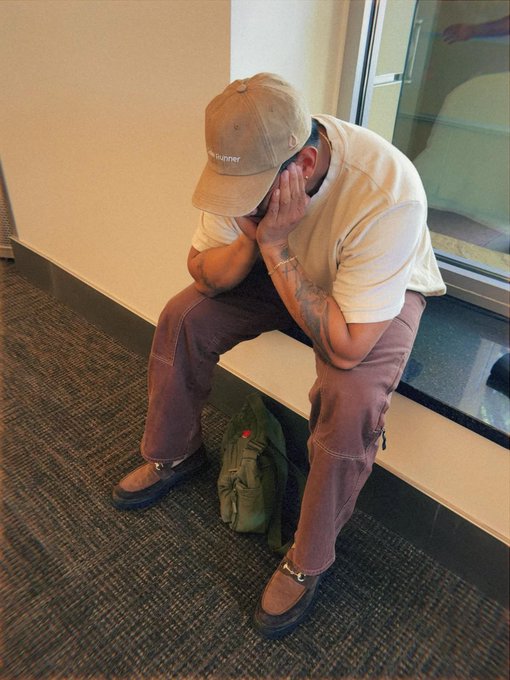 A man wearing a tan cap, white t-shirt, and brown pants sits hunched over on a ledge with his face in his hands, a green bag on the floor beside him.