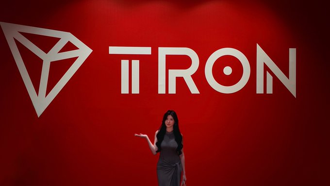 A woman in a black dress gestures toward a large TRON logo displayed on a red backdrop in Singapore. The image visually reinforces TRON's branding and coincides with promotional stats highlighting the blockchain's 407 million total accounts and massive $30 trillion cumulative transfer volume.