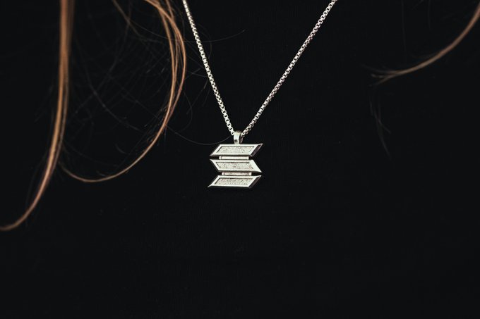 A silver pendant necklace with three stacked banners hanging from a thin chain, displayed against a dark background with strands of hair visible.
