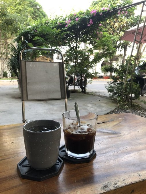 A rustic outdoor café scene with two glasses of iced coffee on a table, set under a trellis draped in pink flowering vines.