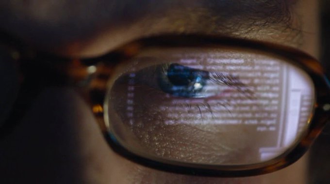 A close-up shot of a person's eye reflected in a tortoiseshell eyeglass lens, showing lines of code scrolling across the glass surface. The image visually reinforces the post's theme of decentralized AI oversight by depicting human observation of code, symbolizing transparency and scrutiny in machine-driven decision-making.