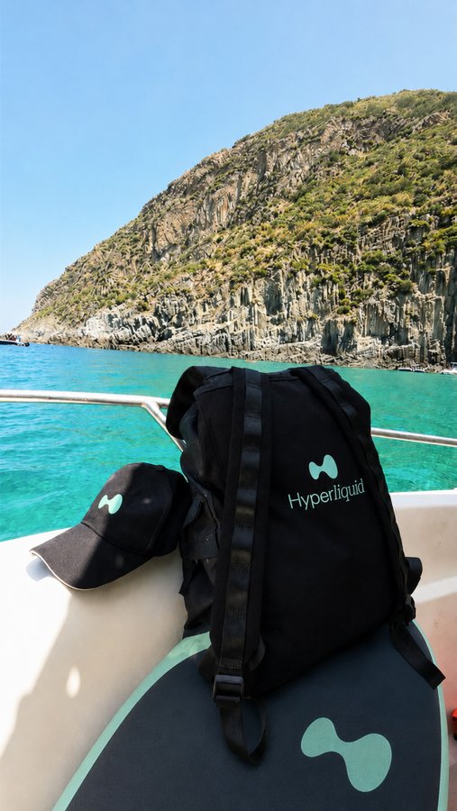 A black backpack and matching cap branded with "Hyperliquid" and its logo sit on a boat seat, with turquoise water and a rocky coastline in the background.