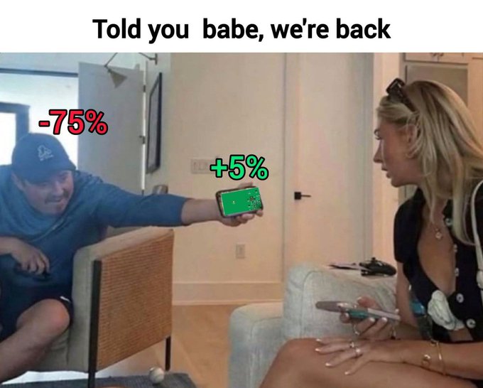 The image shows a man proudly showing his phone to a woman, with the caption "Told you babe, we're back," displaying "+5%" on his phone (representing a small gain) while labeled "-75%" (indicating his overall portfolio loss), humorously depicting how crypto holders cling to minor green candles while still sitting on massive losses.