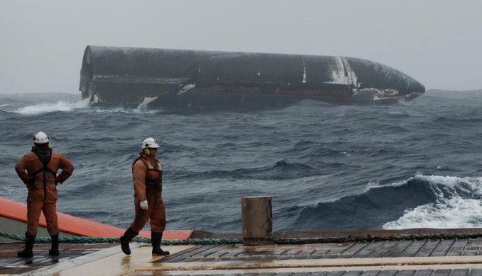 Two workers in orange protective gear watch rough seas aboard a ship as a large capsized vessel lies on its side in the background. The image accompanies the post “Starship Odyssey,” fitting its maritime disaster or shipwreck theme.