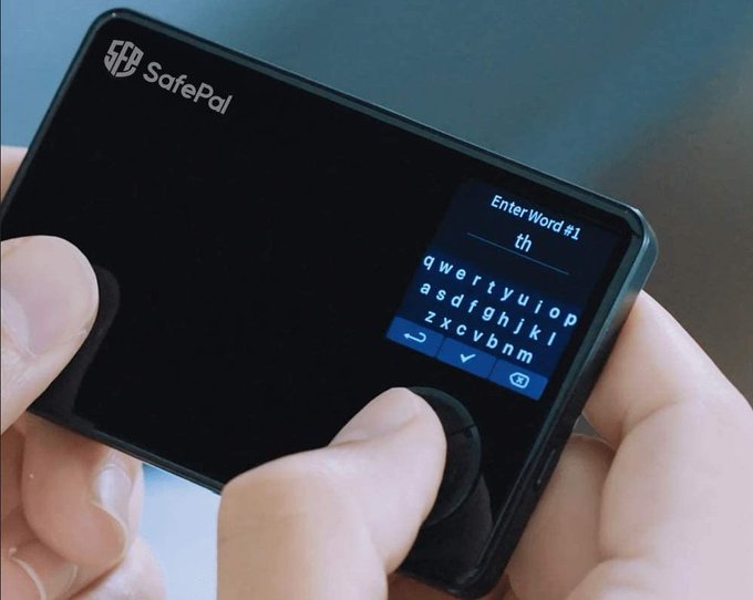 A close-up photo of hands operating a SafePal hardware cryptocurrency wallet, entering the first word of a recovery seed phrase on its small touchscreen with a QWERTY keyboard.