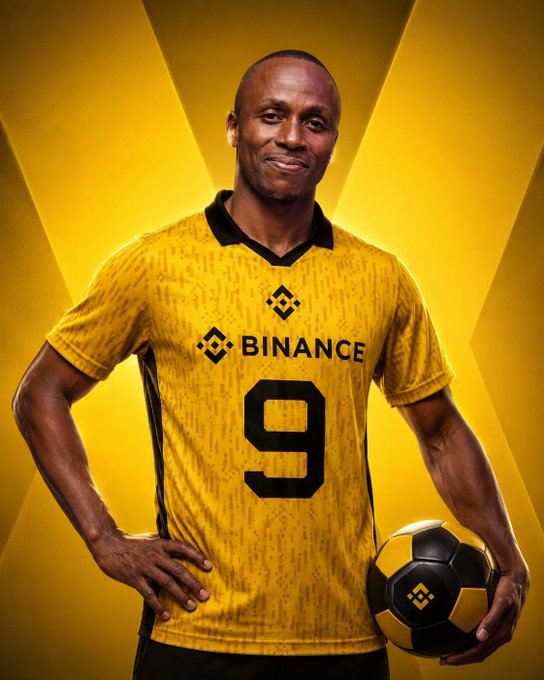 A man wearing a yellow Binance jersey with the number 9, holding a soccer ball featuring the Binance logo against a yellow geometric background.