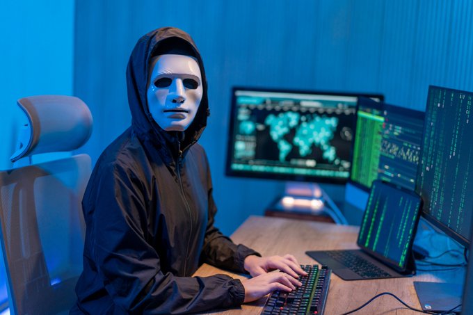A person wearing a white mask and dark hoodie sits at a desk with multiple monitors displaying world maps and code, evoking a stereotypical hacker image.