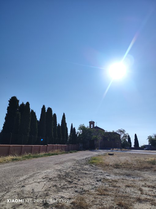 Outdoor photograph taken with a Xiaomi 12T Pro (watermark 'SACRA97') showing a dirt road lined with tall cypress trees, a small chapel with a bell tower, and a bright sun creating lens flare in a clear blue sky.