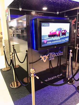 A Bitcoin ATM kiosk branded 'Bitcoin ATM' by Ferris, displaying an advertisement featuring a pink convertible and the website www.ferrisecars.com, surrounded by stanchions.