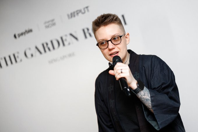A young man with glasses and short hair, wearing a black jacket, speaks into a microphone in front of a backdrop that includes the text 'THE GARDEN ROUTE'.