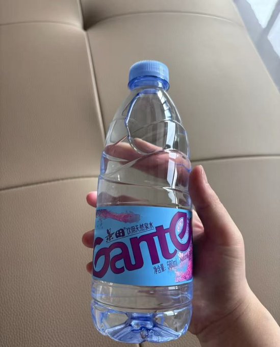 A hand holds a 550ml bottle of 景田 (Gante) drinking natural mineral water, with the label visible in light blue and pink. The image humorously plays into the post about a colleague using the water bottle as a playful prop for an extended "handshake" bit referencing Justin Sun.