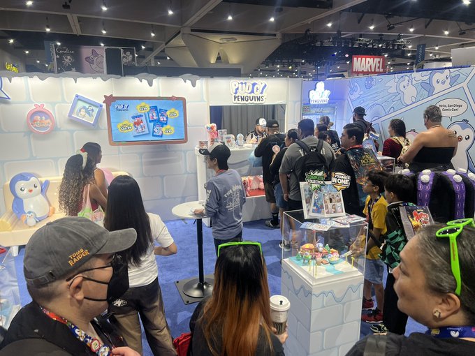 A crowded convention floor scene showing attendees at various booths, including "Pudgy Penguins" and "Marvel," with people browsing merchandise, watching displays, and interacting in a large exhibition hall.