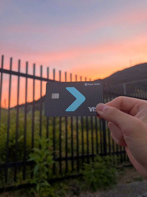 A hand holds up a dark "Bitget Wallet" Visa card against a stunning sunset with pink and orange hues, framed by a black metal fence and silhouetted hills. The image pairs perfectly with the caption "You can't buy sunsets (for everything else there's the Bitget Wallet Card)," visually emphasizing the message that while some things are priceless, the crypto card handles all other purchases.