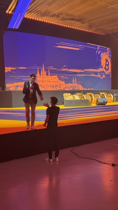 The image shows a group of people standing near a stage with a large screen displaying an advertisement for Bitcoin. A young man stands at the edge of the stage, holding his hand up as if he has just received his first 2,000 sats ( Bitcoins). Another person is seen talking on their cell phone in the background. The scene suggests that they are attending an event or gathering related to Bitcoin and its adoption.