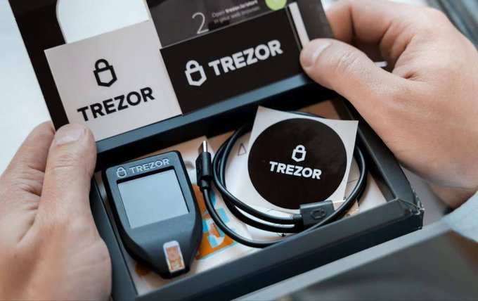 Hands unboxing a Trezor hardware cryptocurrency wallet, showing the device, USB cable, instruction card, and branded packaging materials.