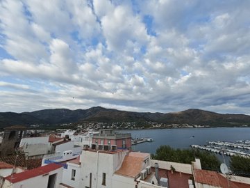 An elevated view of a coastal town or city under a dramatic, cloudy sky, looking out over red-roofed buildings towards a harbor and lush green mountains.