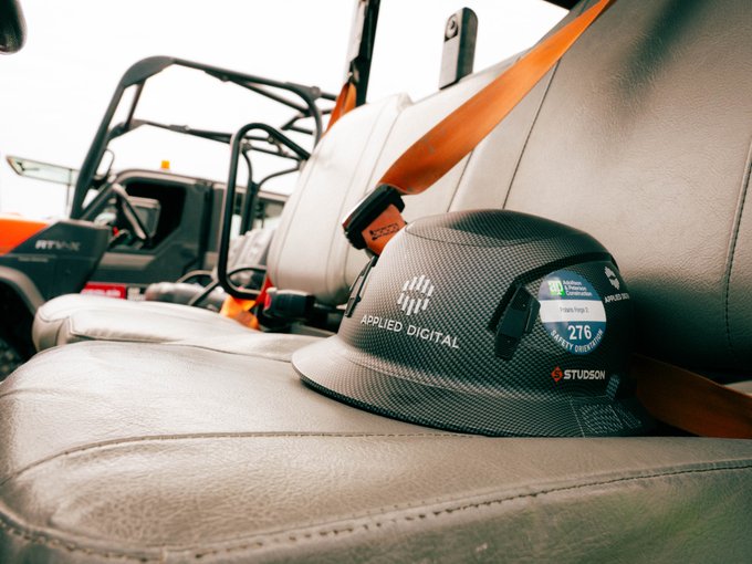 A black carbon-fiber "APPLIED DIGITAL" hard hat with several stickers (including "276", "amazon", and "STUDSON") resting on a tan leather seat of an orange construction vehicle or ATV, with an orange seatbelt visible.