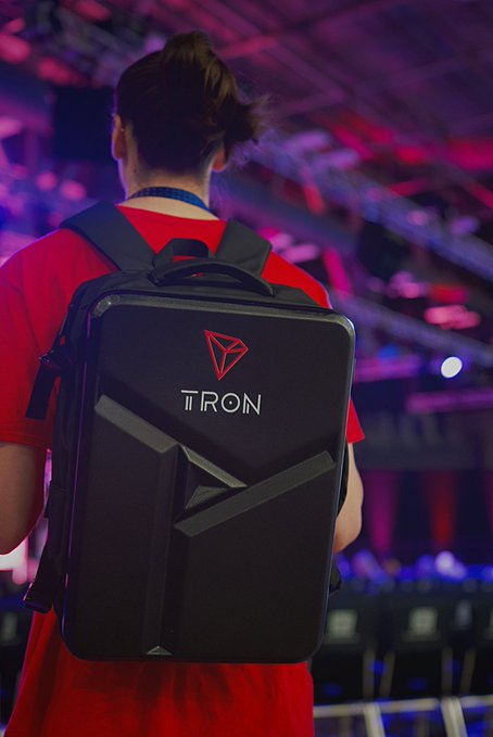 A person with a top knot bun wearing a red shirt and a black backpack branded with the 'TRON' logo stands in a venue lit with purple and blue lighting.